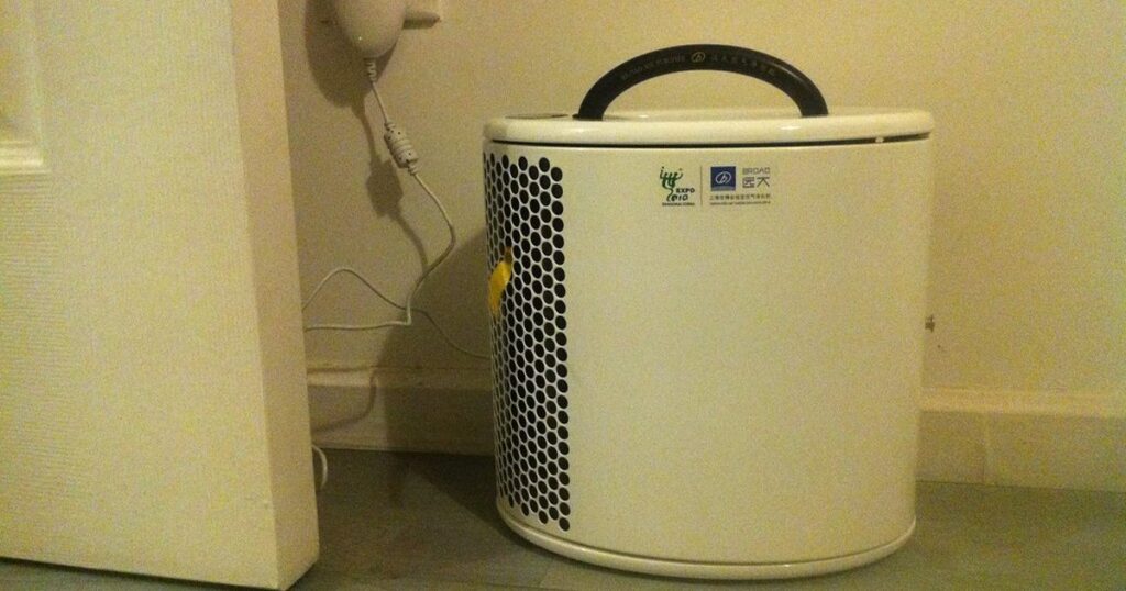 A white freestanding home air purifier with a slotted intake grille standing on a floor beside a wall in a room.
