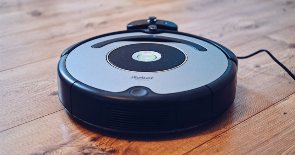 A round black robot vacuum resting on a hard floor near the edge of a light area rug.