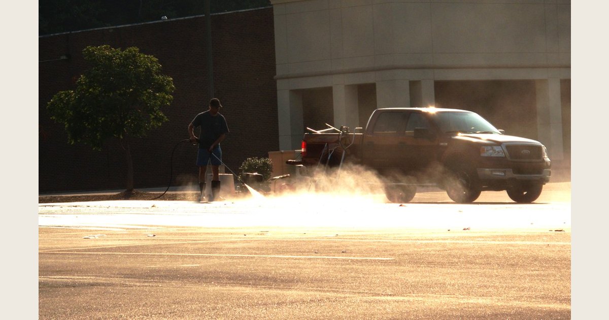 A person operates a pressure washer near a parked vehicle, with water spraying across the pavement.
